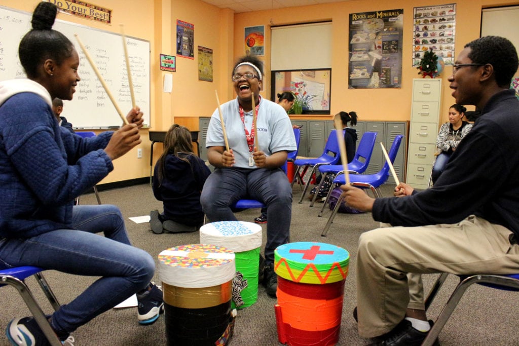 Three laughing students playing their bucket drums in a classroom