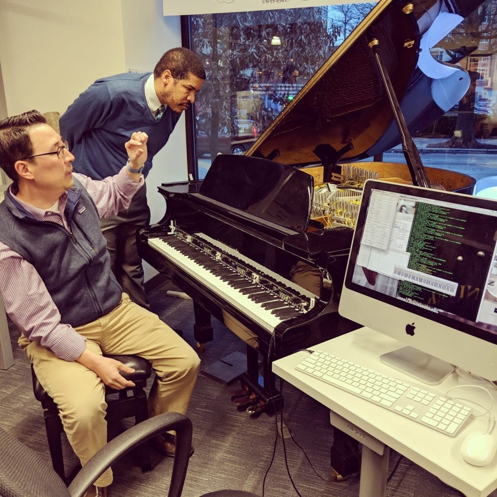 ExCITe Center director Youngmoo Kim demonstrates the ExCITe Center’s magnetic resonator piano. Youngmoo sits on the piano bench, while composer Jay Fluellen looks into the open lid of a baby grand piano. There is a computer monitor and keyboard next to the piano keyboard.