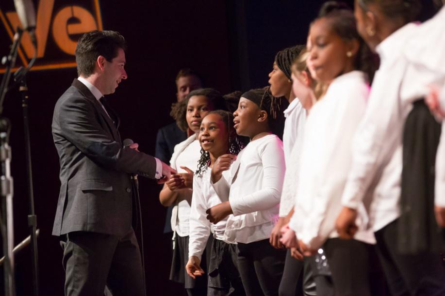 Paul Smith (left) of VOCES8 talks on stage with elementary students from C.W. Henry School. About 8 students are standing in a line, wearing matching white shirts, and one girl is pointing to herself with excitement as Paul gestures toward her.