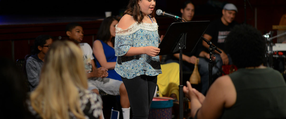 Samantha, a student from The Center for Grieving Children, stands in front of other students attending the Percussion Studio as she recites her poem into a microphone.