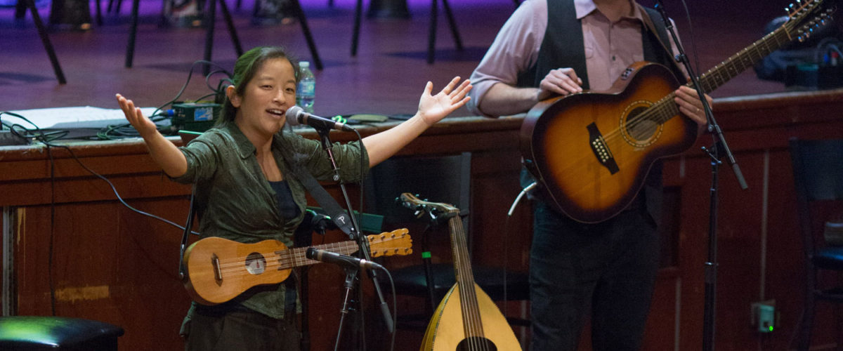 Elena Moon Park holds a jarana and opens her hands wide. Next to her is Ami Yares with his guitar and oud.